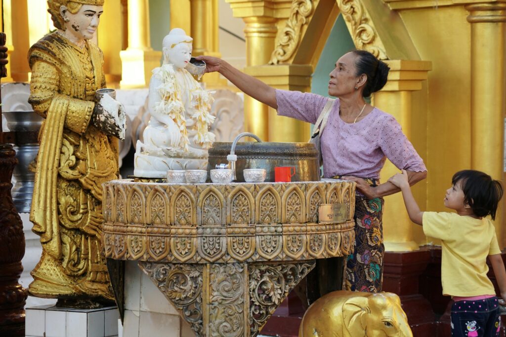 On the first day of the Songkran Festival, a Thai grandma visits a beautiful golden temple, where she pours water scented with fragrant flowers over the shoulder of a white statue of Buddha. Her grandchild clings to her arm and watches this sacred ritual. 